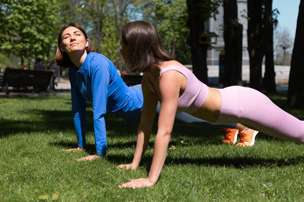 Dos mujeres bonitas en ropa deportiva sobre el césped en el parque en un día soleado haciendo planta de entrenamiento se apoyan mutuamente emociones felices