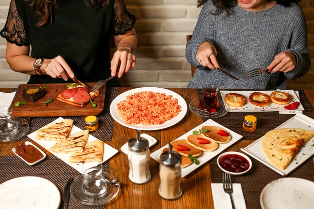 Dos mujeres desayunan con tostadas, crepes, panqueques, mermelada y huevos revueltos