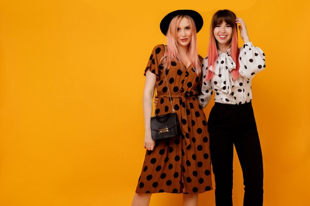 Dos mujeres elegantes con estilo en vestidos posando sobre pared amarilla