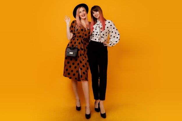 Dos mujeres elegantes con estilo en vestidos posando sobre pared amarilla