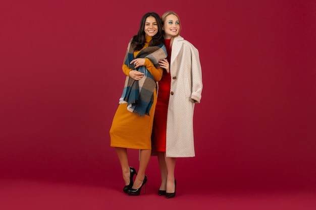 Dos mujeres elegantes en vestido de moda otoño invierno y abrigo posando aislado en la pared roja