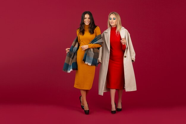 Dos mujeres elegantes en vestido de moda otoño invierno y abrigo posando aislado en la pared roja