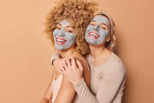 Dos mujeres felices se abrazan y se paran de lado contra el fondo marrón aplican una máscara de arcilla de belleza en las caras para eliminar las líneas finas y los poros sonríen ampliamente disfrutan del tiempo para ellas mismas Concepto de cuidado de la piel