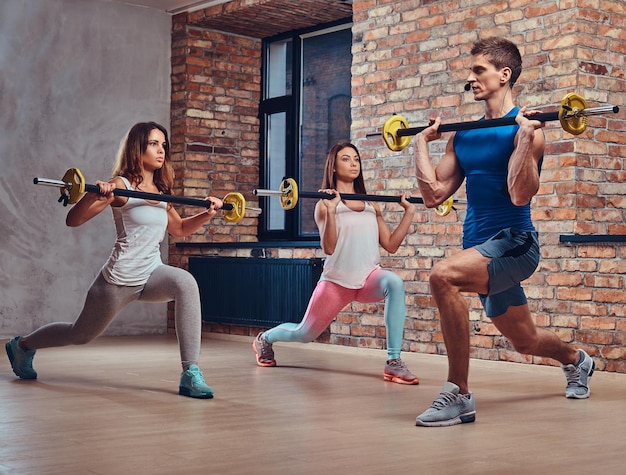 Dos mujeres fitness y un entrenador, estirando sentadillas con pesas en un club con interior de loft.