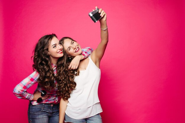 Dos mujeres hermosas y de moda vestidas con ropa elegante que pasan tiempo juntas haciendo fotos en la cámara Novias positivas posando sonriendo y tomando autorretrato Fondo rosa