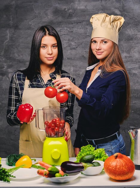 Dos mujeres jóvenes atractivas preparando jugo de vegetales veganos con licuadora.