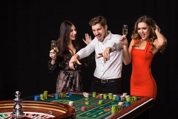 Dos mujeres jóvenes y un hombre detrás de la mesa de la ruleta sobre fondo negro. Jugadores de emociones