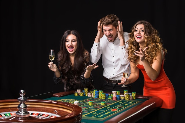 Dos mujeres jóvenes y un hombre detrás de la mesa de la ruleta sobre fondo negro. Jugadores de emociones