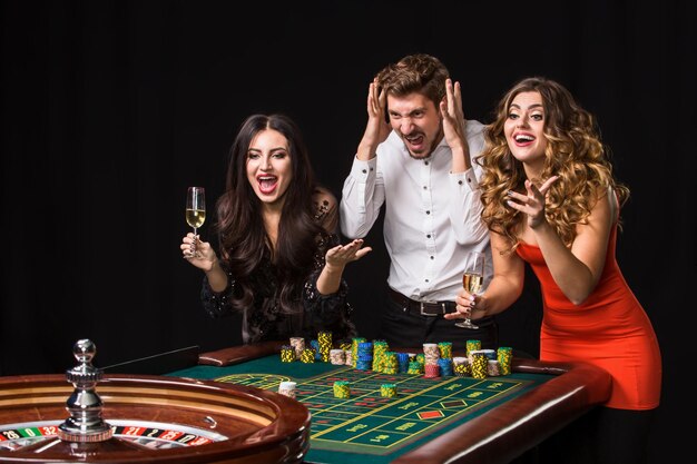 Dos mujeres jóvenes y un hombre detrás de la mesa de la ruleta sobre fondo negro. Jugadores de emociones