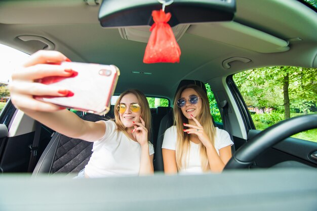 Dos mujeres jóvenes se juntan en un viaje en automóvil conduciendo el automóvil y tomando selfie