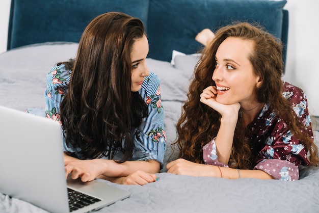 Dos mujeres jóvenes que miran uno a con la computadora portátil en cama