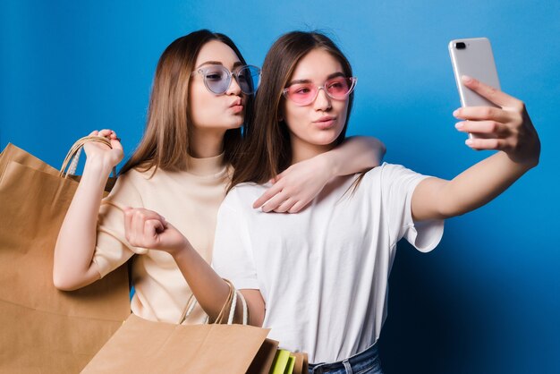 Dos mujeres jóvenes toman selfie en el teléfono con bolsas de papel de colores aislados en la pared azul. Concepto de venta en tienda.