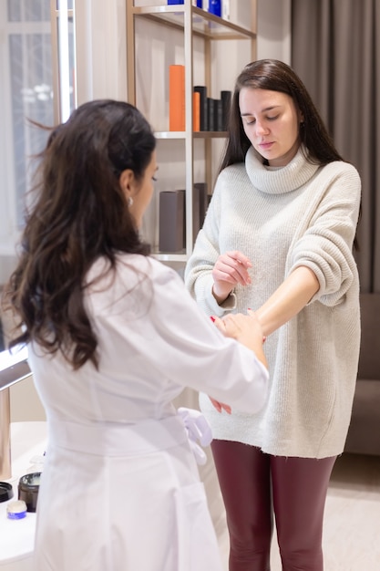 Dos mujeres, un médico esteticista y un cliente, se paran en el espejo, para una consulta, el médico aplica crema en las manos de la mujer y hace un ligero masaje.