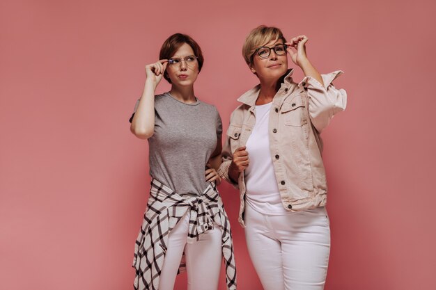 Dos mujeres de moda de pelo corto con gafas en pantalones pitillo blancos y camisetas frescas posando sobre fondo rosa aislado.