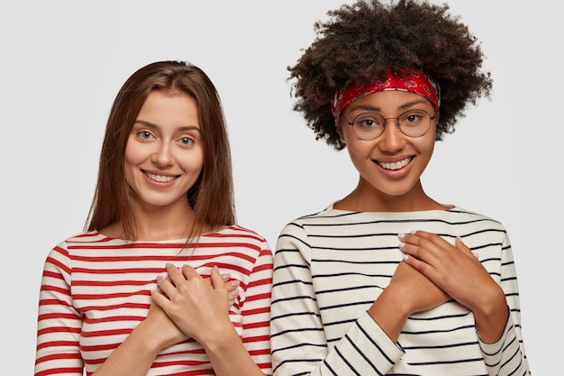 Foto gratuita dos mujeres multiétnicas felices y encantadas se dan la mano en el pecho, sonríen con alegría y recuerdan el gran momento, aprecian el apoyo de alguien, se sienten agradecidos, usan jerséis de rayas, aislados sobre una pared blanca