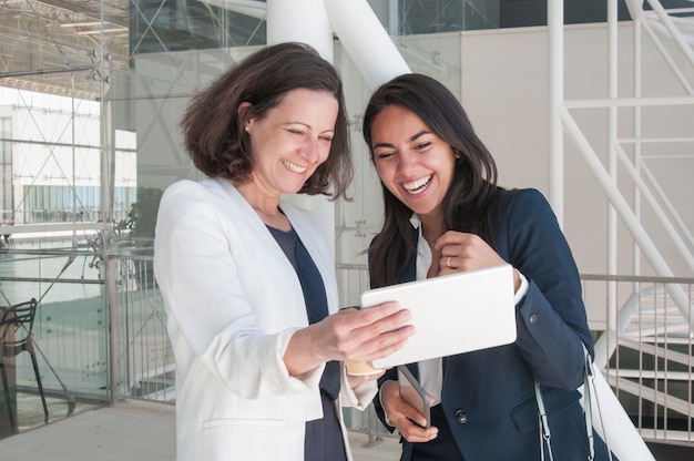 Dos mujeres de negocios sonrientes que usan la tableta en pasillo de la oficina