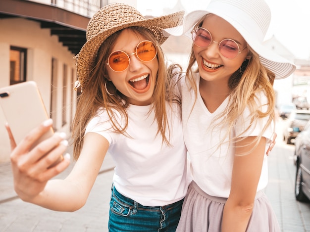 Dos mujeres rubias sonrientes jovenes del inconformista en la camiseta blanca del verano. chicas tomando fotos de autorretrato autofoto en el teléfono inteligente. modelos posando en el fondo de la calle. la mujer muestra emociones positivas