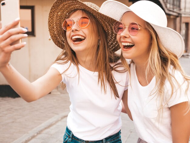 Dos mujeres rubias sonrientes jovenes del inconformista en la camiseta blanca del verano. Chicas tomando fotos de autorretrato en el teléfono inteligente. Modelos posando en el fondo de la calle. La mujer muestra lengua y emociones positivas.