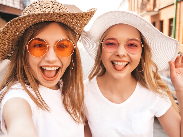 Dos mujeres rubias sonrientes jovenes del inconformista en la camiseta blanca del verano. Chicas tomando fotos de autorretrato en el teléfono inteligente. Modelos posando en el fondo de la calle. La mujer muestra lengua y emociones positivas.