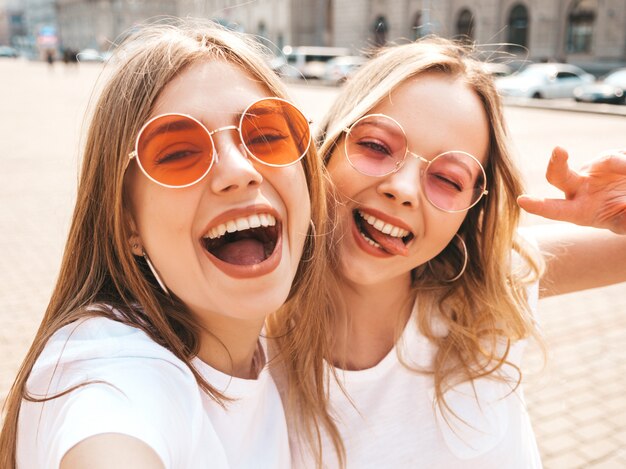 Dos mujeres rubias sonrientes jovenes del inconformista en ropa blanca de la camiseta del verano. Chicas tomando fotos de autorretrato en el teléfono inteligente. Modelos posando en la calle. La mujer muestra el signo de la paz y la lengua.