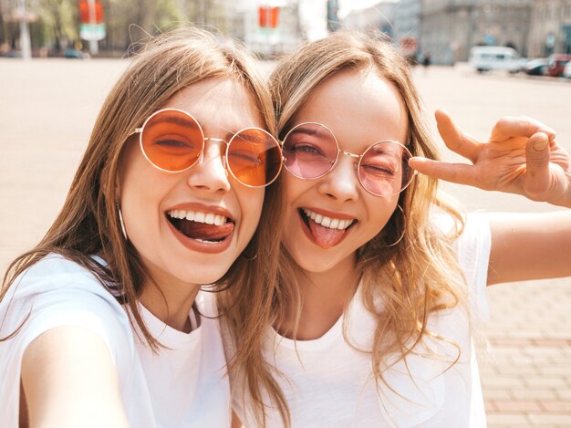 Dos mujeres rubias sonrientes jovenes del inconformista en ropa blanca de la camiseta del verano. Chicas tomando fotos de autorretrato en el teléfono inteligente. Modelos posando en la calle. Mujer positiva mostrando su lengua