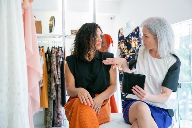 Dos mujeres sentadas juntas y usando tableta, discutiendo ropa y compras en la tienda de moda. Vista frontal. Consumismo o concepto de compras