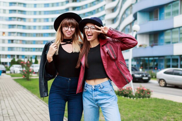 Dos mujeres sonrientes despreocupadas posando en la ciudad moderna. Vistiendo gorro de lana, chaqueta de cuero y jeans.