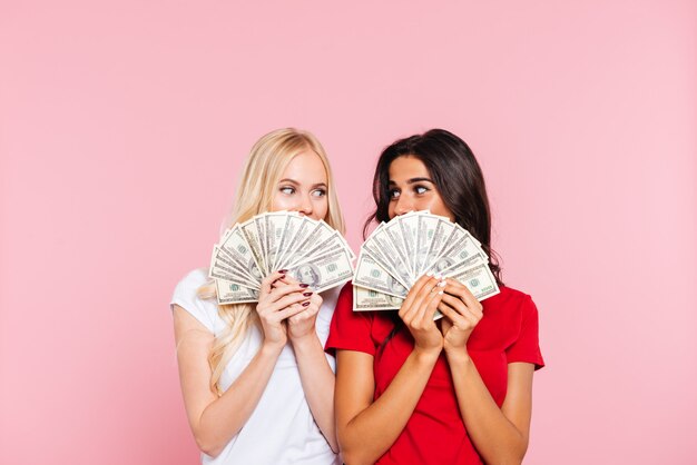 Dos mujeres sonrientes escondiéndose detrás del dinero y mirándose sobre rosa
