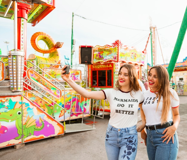 Dos mujeres sonrientes que toman selfie en teléfono inteligente en el parque de atracciones