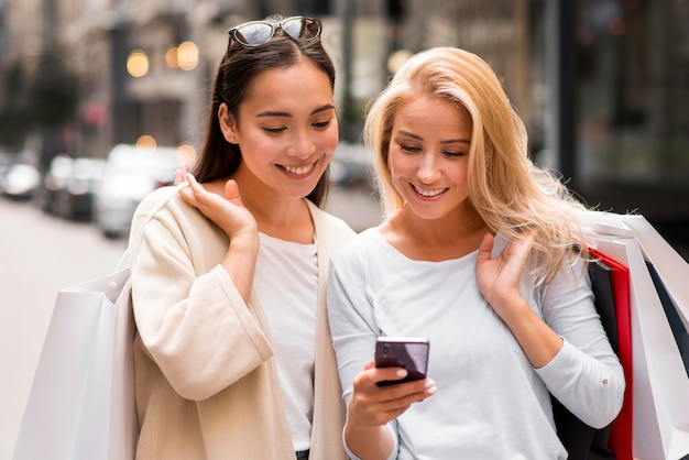 Foto gratuita dos mujeres sosteniendo bolsas de compras y mirando smartphone