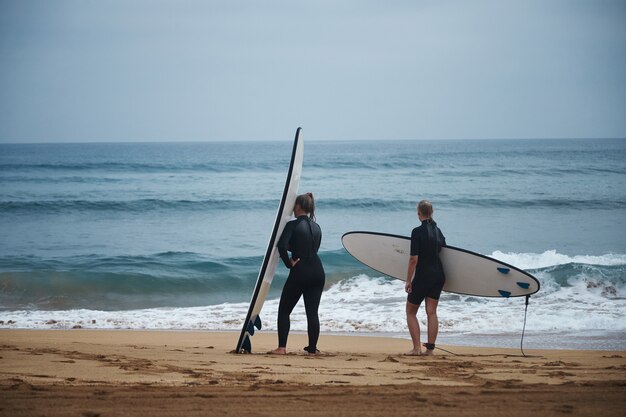 Dos mujeres en trajes de neopreno con tablas de surf se preparan para ir al agua en un día fresco de verano