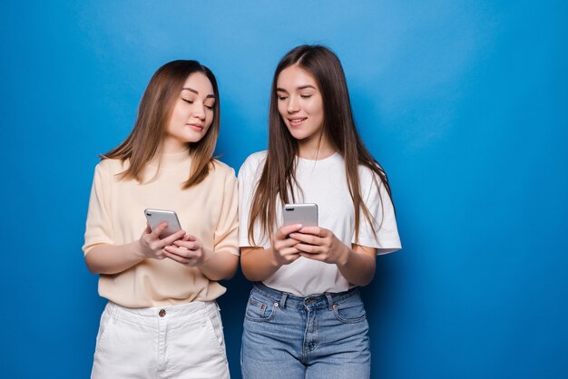 Dos mujeres de verano multiétnicas vistiendo expresando emoción o sorpresa mientras ambos usan teléfonos celulares sobre la pared azul