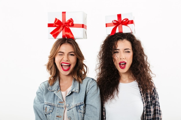 Dos niñas felices con regalos en la cabeza mientras guiña un ojo a la cámara sobre la pared blanca