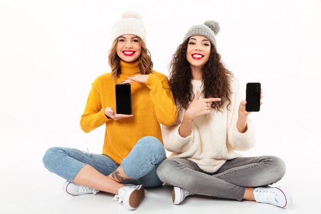 Dos niñas felices con suéteres y sombreros sentados juntos en el suelo mientras muestran pantallas en blanco de teléfonos inteligentes sobre la pared blanca