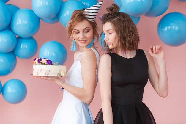 Dos niñas lindas adolescentes de la moda que sostienen la torta poner crema blanca del feliz cumpleaños.