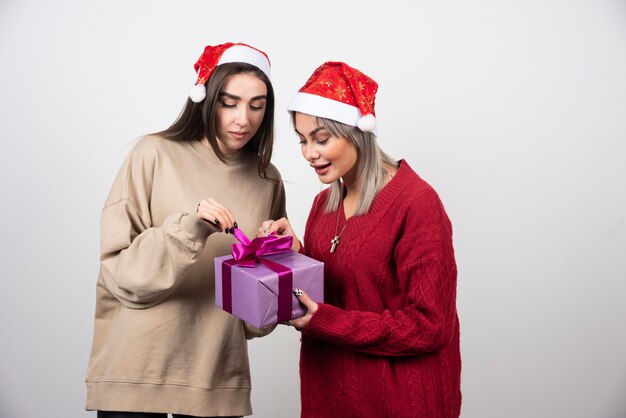 Dos niñas sonrientes con gorro de Papá Noel que envuelven un regalo de Navidad festivo.