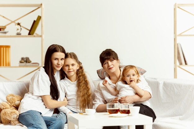 Dos niñas, su atractiva madre joven y su encantadora abuela sentados en el sofá y pasar tiempo juntos en casa. Generación de mujeres. Día Internacional de la Mujer. Feliz día de la madre.