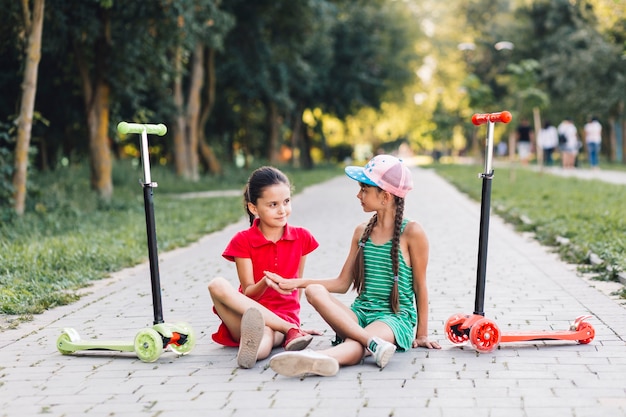 Dos niñas se tocan la mano mientras están sentadas en la pasarela con sus scooters de empuje