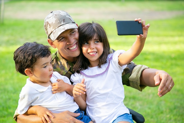 Dos niños alegres sentados en el regazo de los papás y tomando selfie en el celular. Hombre militar discapacitado caminando con niños en el parque. Veterano de guerra o concepto de discapacidad