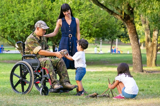 Dos niños arreglando madera para fogatas al aire libre cerca de mamá y papá militar discapacitado en silla de ruedas. Niño mostrando registro al padre. Veterano discapacitado o concepto de familia al aire libre