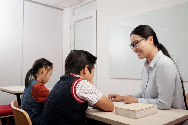 Dos niños asiáticos sentados en el aula, y profesor sonriente con gafas hablando con boy