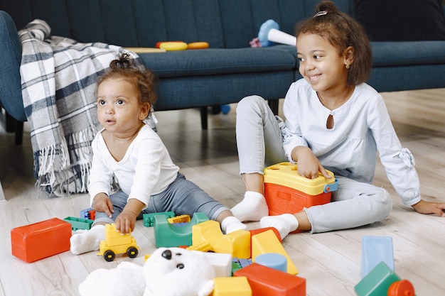 Dos niños bonitos se sientan en el suelo y juegan con juguetes cerca del sofá. Hermanas afroamericanas jugando en casa.