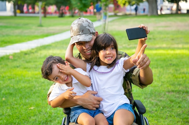 Dos niños felices sentados en el regazo de los papás y tomando selfie en celular. Hombre militar discapacitado caminando con niños en el parque. Veterano de guerra o concepto de discapacidad