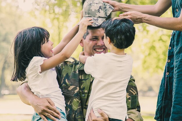 Dos niños felices y su mamá se encuentran y abrazan a papá militar en uniforme de camuflaje al aire libre