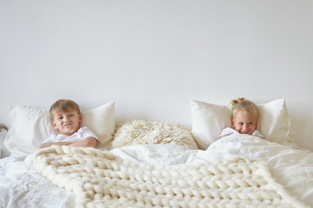 Dos niños lindos relajándose en el dormitorio. Filmación en interiores de un adolescente en pijama acostado en la cama con su hermano pequeño rubio en el otro lado, con miradas divertidas. Concepto de infancia, niños y familia.