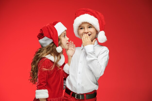 Dos niños y niñas felices con sombreros de santa claus con cajas de regalo en el estudio rojo