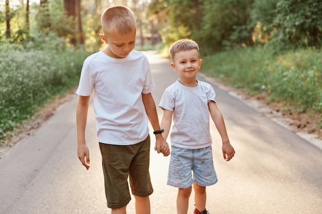 Dos niños pequeños con camisetas blancas y pantalones cortos que van juntos y tomados de la mano en el parque de verano, hermanos caminando al aire libre, expresando emociones positivas.