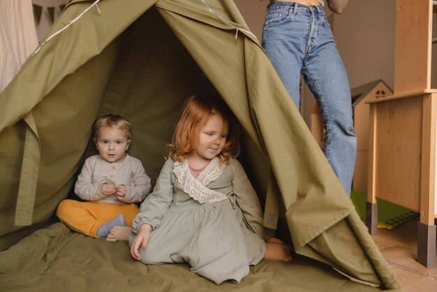 Dos niños pequeños rubios de piel clara con una chica pelirroja visten ropa informal sentados en una tienda de campaña en el interior Concepto de familia