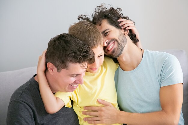 Dos padres guapos felices y su hijo sentados en el sofá juntos y abrazándose. Concepto de familia y paternidad feliz