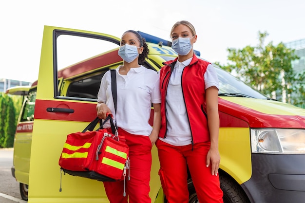 Dos paramédicos de pie junto a la ambulancia Dos enfermeras de emergencia con mascarilla Paramédico con una bolsa de trauma médico